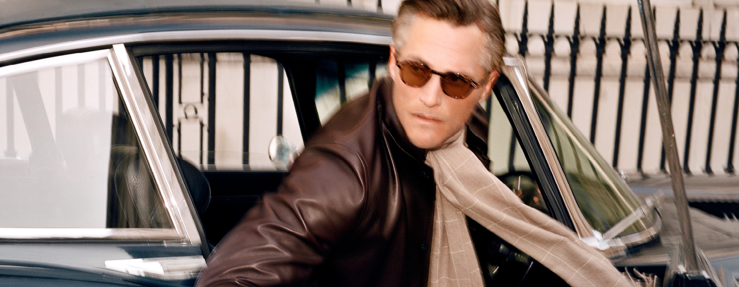 A stylish man wearing sunglasses, a brown leather jacket, and a patterned scarf is stepping out of a vintage blue car. The background features a wrought iron fence.