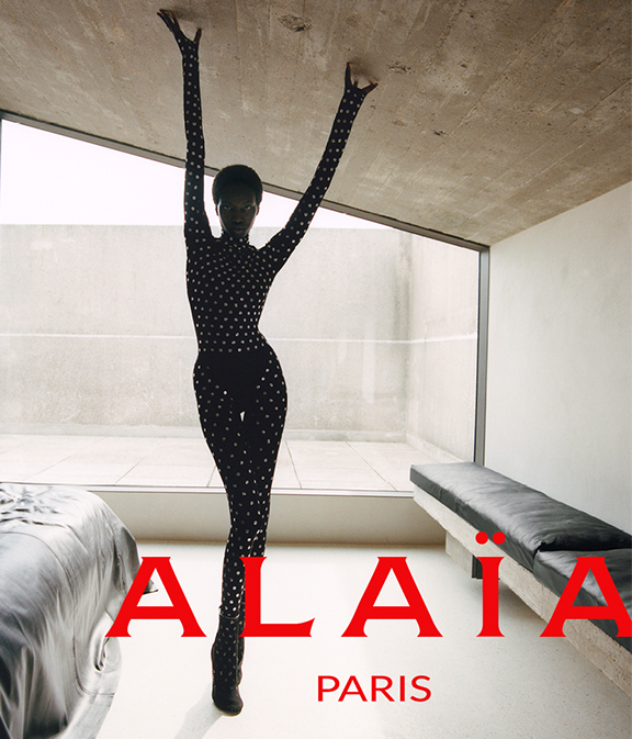 Woman in a bedroom wearing black outfit plus Alaia logo in red