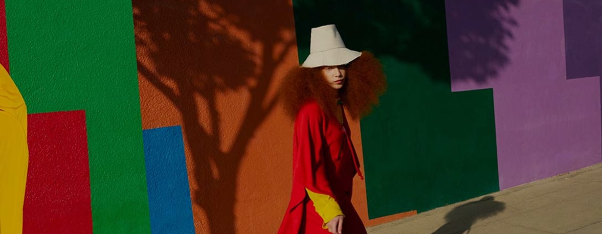 A woman wearing a colourful dress and a hat standing in front of a colourful wall