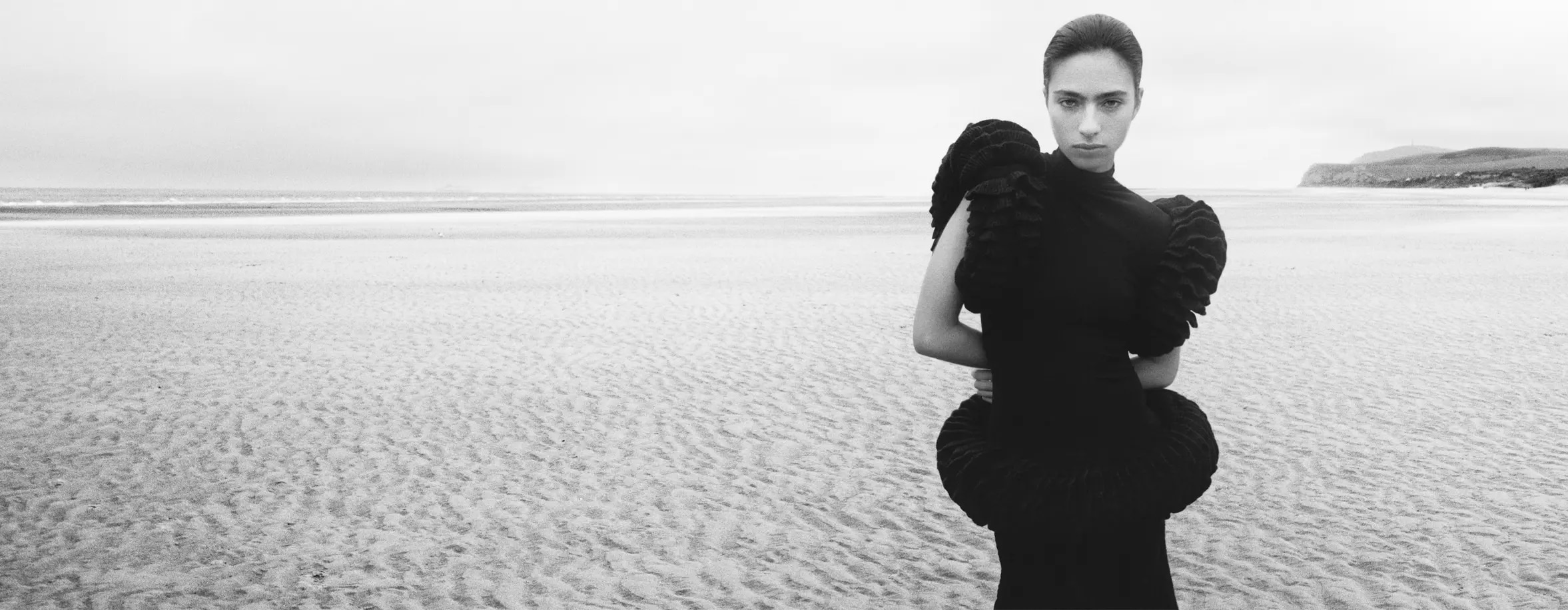 A striking black and white photograph of a woman in a sculptural black dress, standing on a vast, empty beach, creating a sense of drama and isolation.