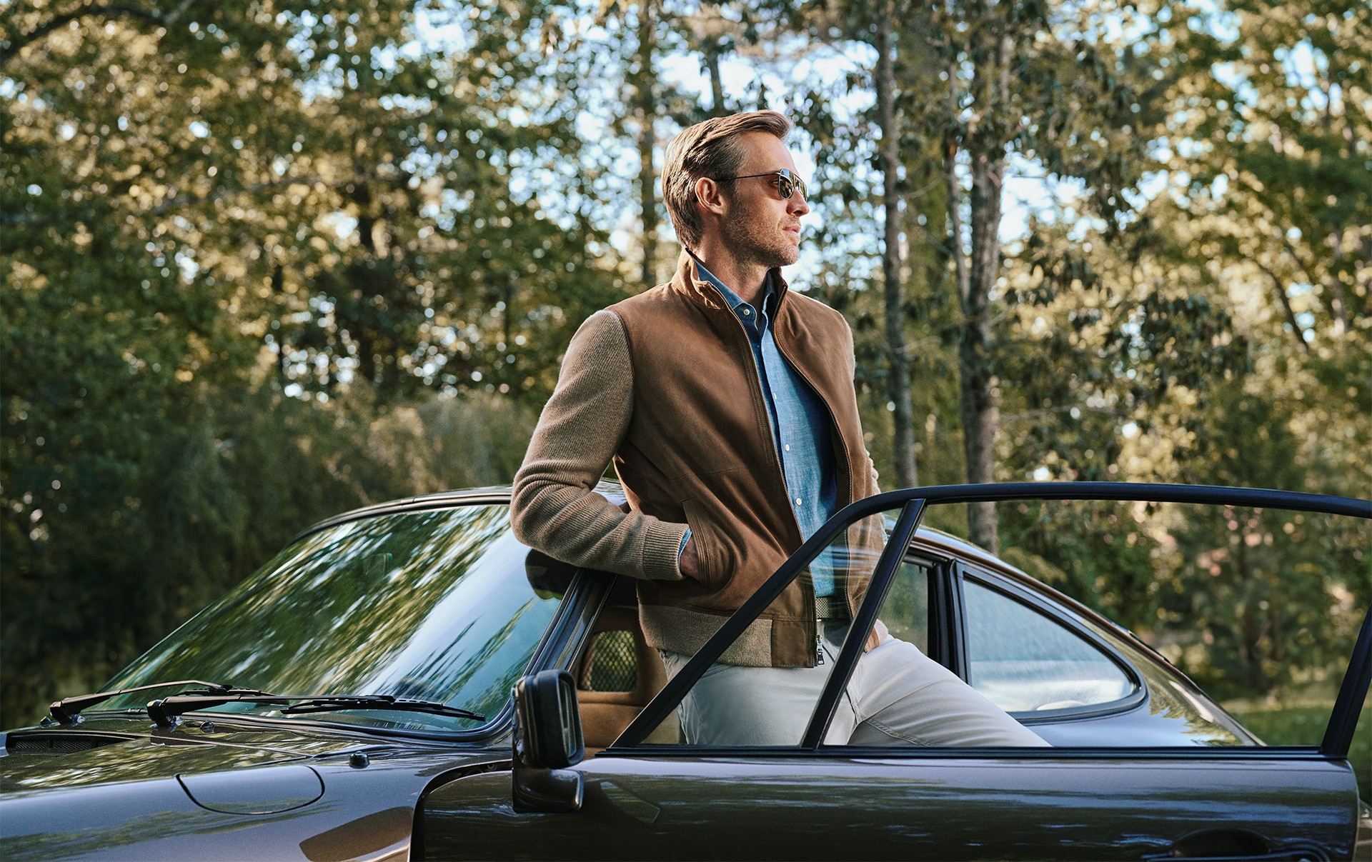 Man in glasses, blue shirt, brown jacket and grey trousers standing by a car